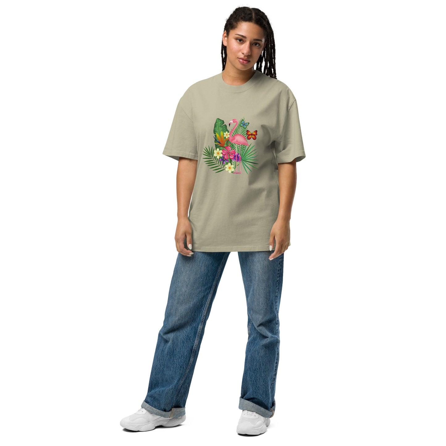 A woman is wearing a eucalyptus oversized t-shirt adorned with a colorful tropical design featuring a pink flamingo surrounded by lush greenery and bright flowers. The playful and vibrant graphic stands out against the tee's dark background. She pairs it with casual blue jeans and white sneakers, presenting a relaxed yet stylish ensemble that evokes a sense of fun and a tropical vibe. Her posture is easygoing, suggesting comfort and a laid-back fashion sense.