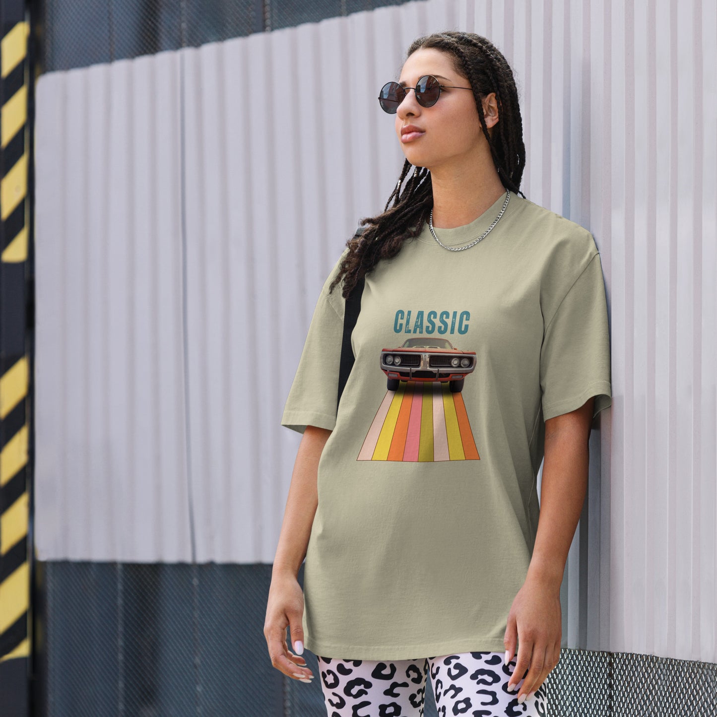 Cool and casual, a woman models a faded eucalyptus 'CLASSIC' graphic tee featuring a vintage car cruising down a retro striped road, and paired with relaxed blue jeans and chic sunglasses.