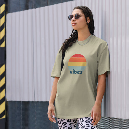 Confident woman showcasing a Women's 'vibes' faded eucalyptus Oversized Tee with a retro-inspired sunset graphic, radiating laid-back, positive vibes in a neutral color, complemented by chic sunglasses. 