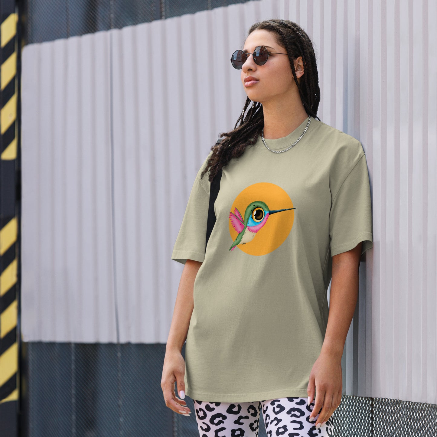 Woman models a faded eucalyptus Oversized Tee with a colorful hummingbird design on a yellow circle background, embodying a lively spirit.