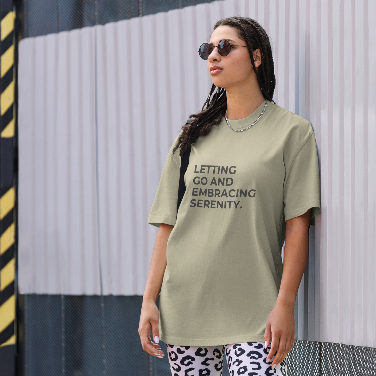 A woman wearing a faded eucalyptus "Letting go and embracing serenity" t-shirt, styled with leopard print leggings and sunglasses, leaning against a corrugated metal wall with a confident expression. The casual yet trendy outfit showcases self-expression and boldness. Perfect for promoting empowering fashion statements.