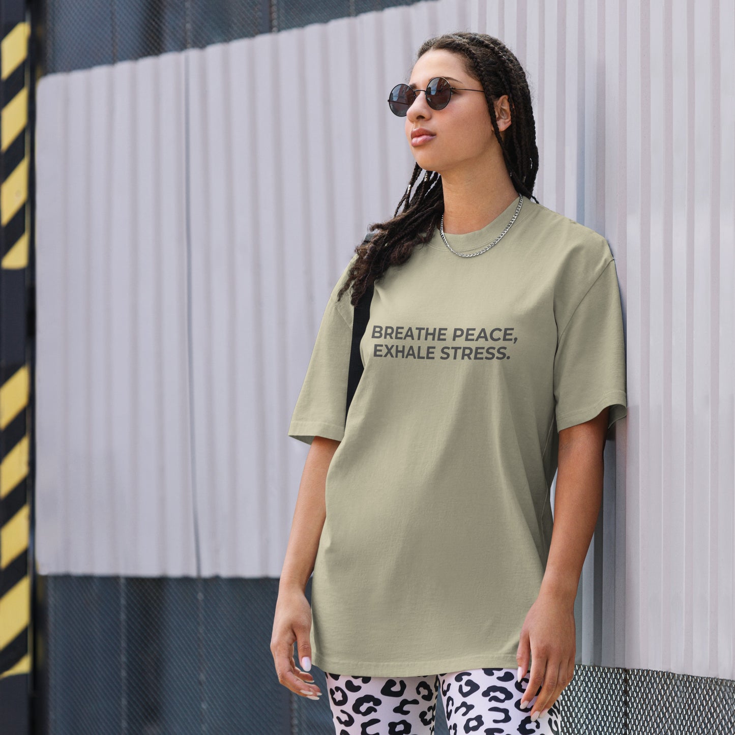 A woman wearing a faded eucalyptus "Breath peace, exhale stress" t-shirt, styled with leopard print leggings and sunglasses, leaning against a corrugated metal wall with a confident expression. The casual yet trendy outfit showcases self-expression and boldness. Perfect for promoting empowering fashion statements.