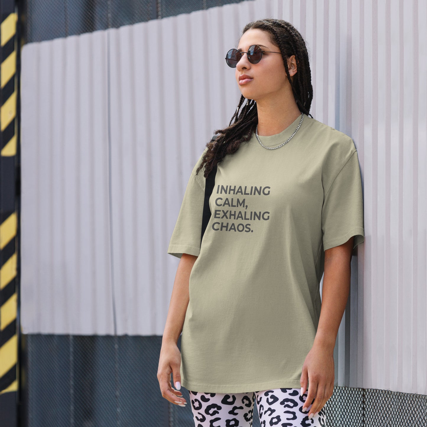 A woman wearing a faded eucalyptus "Inhaling calm, exhaling chaos" t-shirt, styled with leopard print leggings and sunglasses, leaning against a corrugated metal wall with a confident expression. The casual yet trendy outfit showcases self-expression and boldness. Perfect for promoting empowering fashion statements.