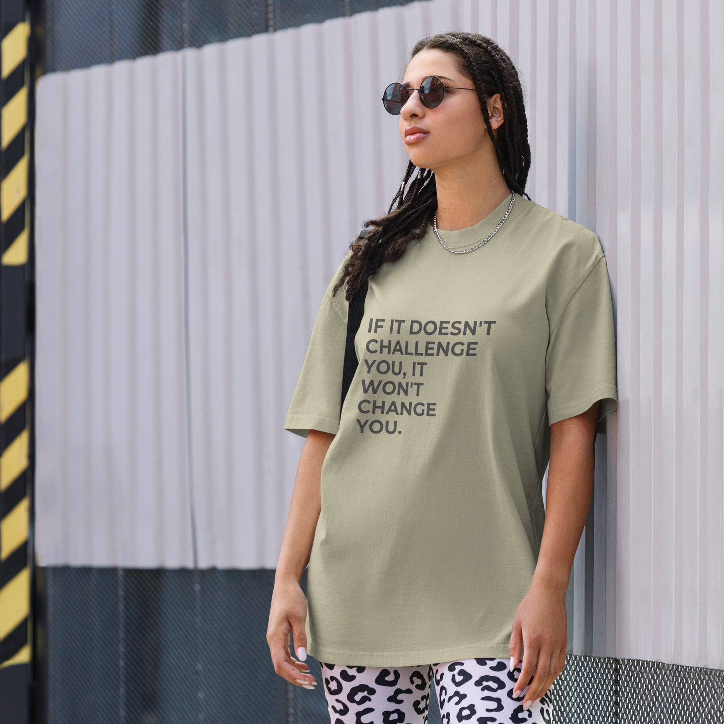 A woman wearing a faded eucalyptus "If it doesn't challenge you, it won'f change you" t-shirt, styled with leopard print leggings and sunglasses, leaning against a corrugated metal wall with a confident expression. The casual yet trendy outfit showcases self-expression and boldness. Perfect for promoting empowering fashion statements.