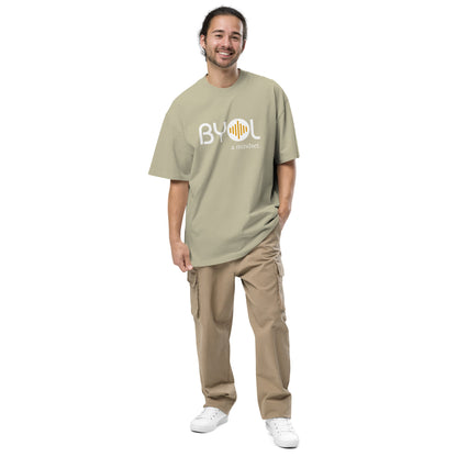 A young man wearing a eucalyptus "BYOL: a mindset" T-shirt, smiling and standing with one hand in his pocket. The T-shirt features the "BYOL" logo in white and yellow on the front and is available in various colors (dark gray, beige, light blue) and sizes (S-3XL). The "be you out loud" logo is displayed at the top.