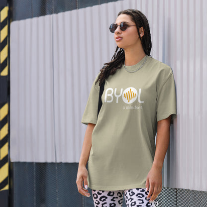 A young woman with long braids wearing sunglasses and a faded eucalyptus "BYOL: a mindset" T-shirt, standing and looking to the side. The T-shirt features the "BYOL" logo in white and yellow on the front and is available in multiple colors (dark gray, beige, light blue, white, plus more) and sizes (S-3XL). The "be you out loud" logo is displayed at the top.