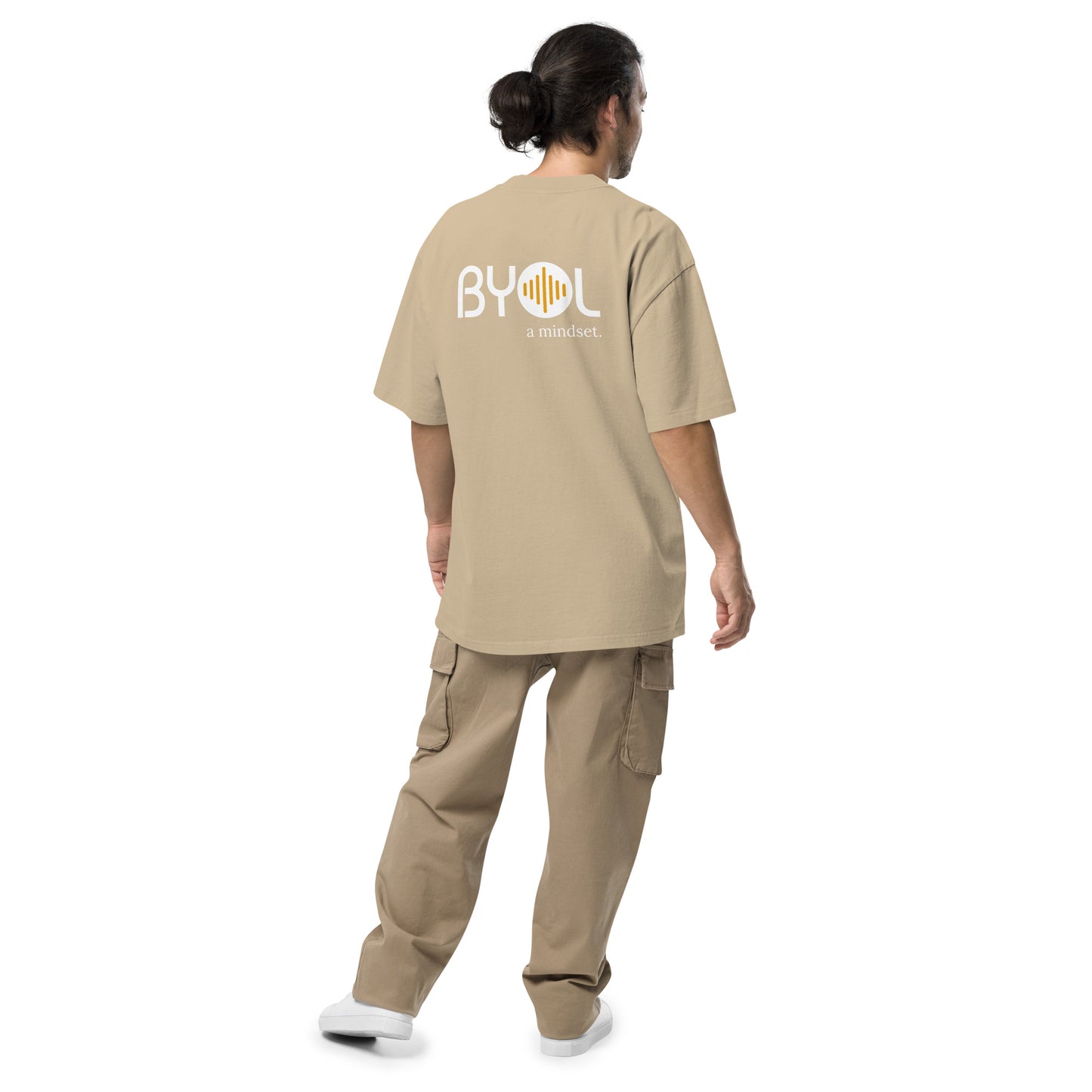 A young man with long hair in a ponytail wearing a khaki "BYOL: a mindset" T-shirt, viewed from the back. The T-shirt features the "BYOL" logo on the back and is available in multiple colors (dark gray, beige, light blue) and sizes (S-3XL). The "be you out loud" logo is displayed at the top.