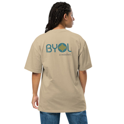 A young woman with long braids wearing a faded khaki "BYOL: a mindset" T-shirt, viewed from the back. The T-shirt features the "BYOL" logo in teal and yellow on the back and is available in multiple colors (dark gray, beige, light blue, white, plus more) and sizes (S-3XL). The "be you out loud" logo is displayed at the top.