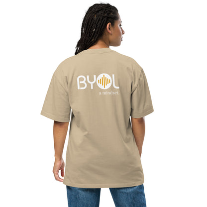 A young woman with long braids wearing a faded khaki "BYOL: a mindset" T-shirt, viewed from the back. The T-shirt features the "BYOL" logo in white and yellow on the back and is available in multiple colors (dark gray, beige, light blue, white, plus more) and sizes (S-3XL). The "be you out loud" logo is displayed at the top.