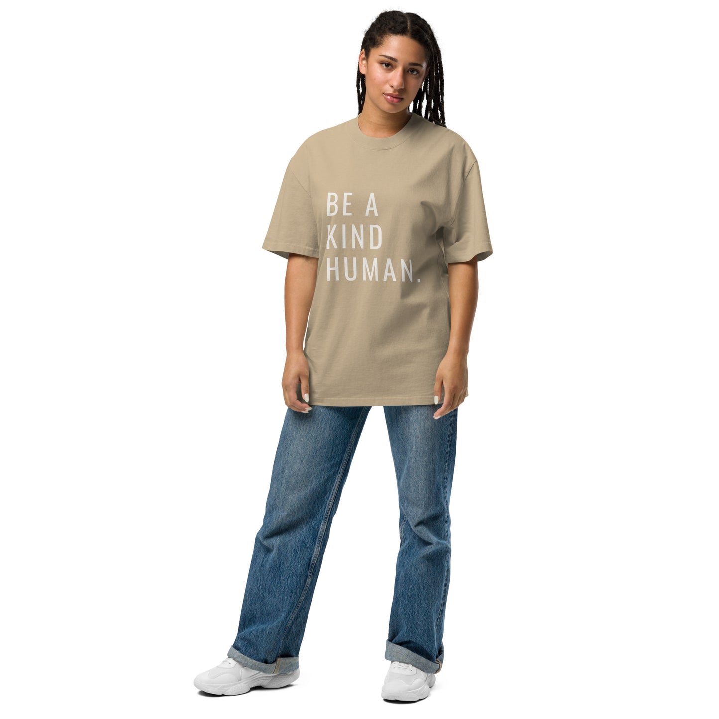 A woman in a casual stance, sporting a faded khaki oversized t-shirt with "BE A KIND HUMAN." in white block letters, complemented by classic blue jeans and white sneakers.