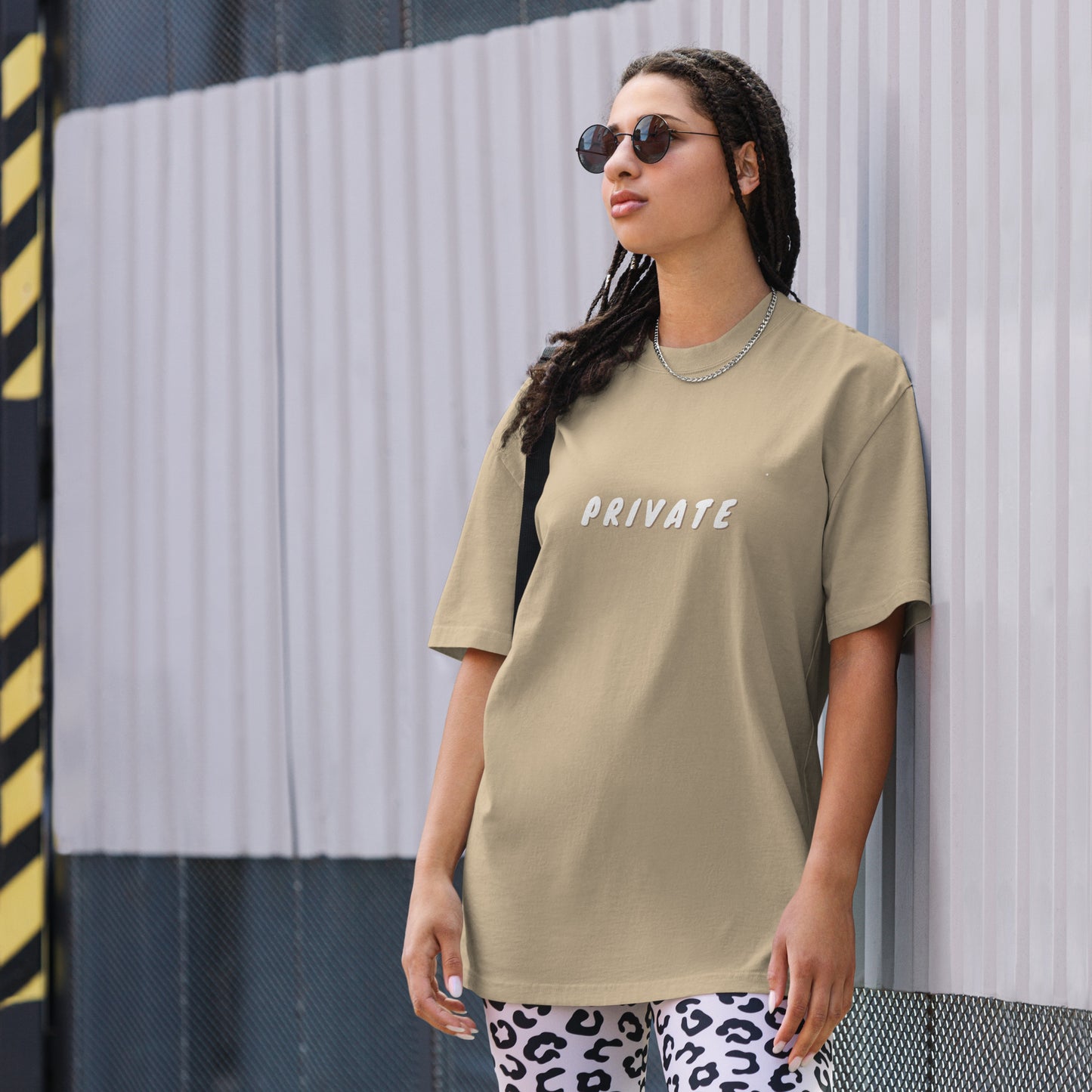 A woman exudes a confident yet understated vibe in an urban setting, wearing a khaki oversized t-shirt with the word "PRIVATE" printed in white across the chest. The simple, bold statement contrasts with her playful white and black leopard print pants. Her style is accessorized with sunglasses and a silver necklace, creating a look that balances a personal statement of discretion with a touch of edgy street fashion.