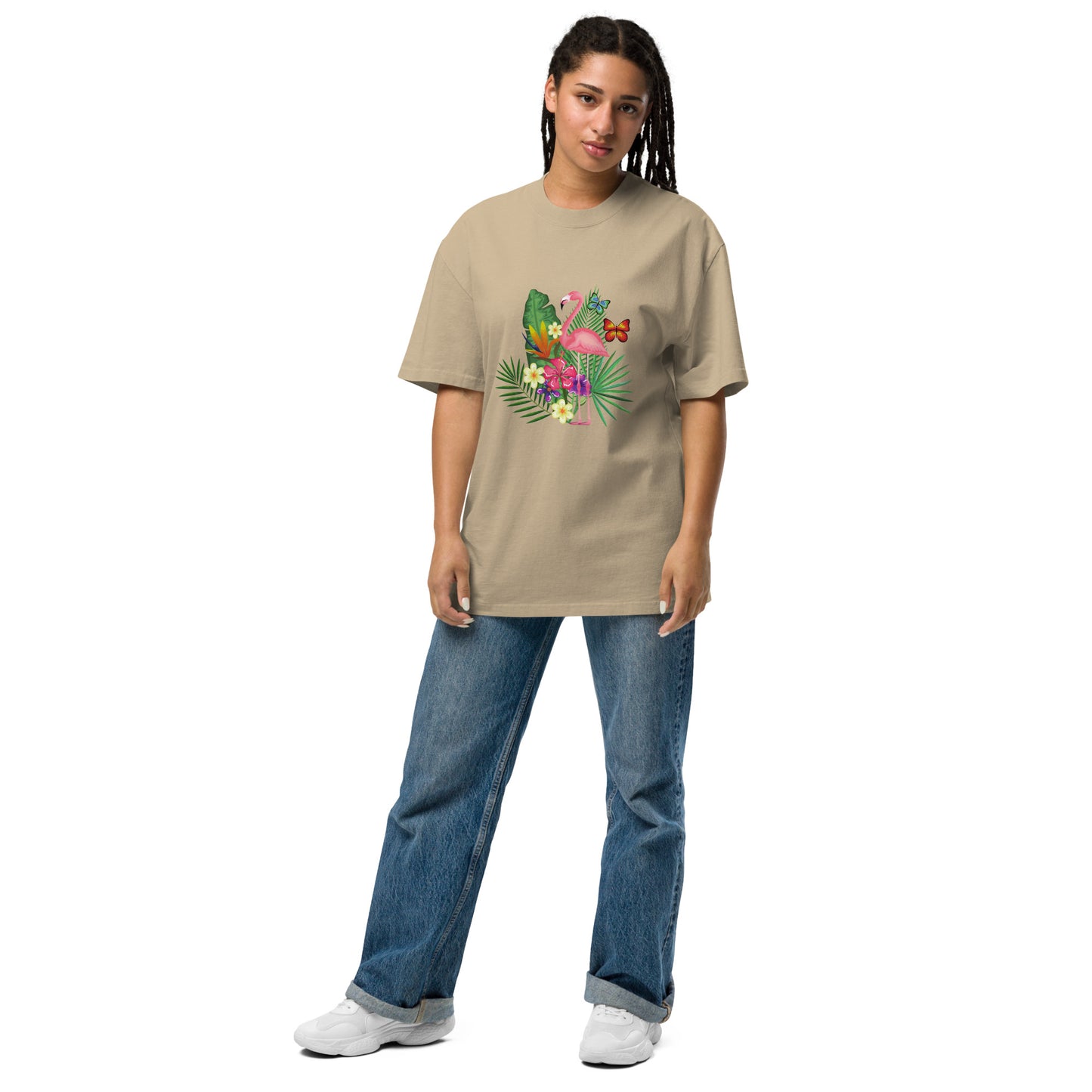 A woman is wearing a khaki oversized t-shirt adorned with a colorful tropical design featuring a pink flamingo surrounded by lush greenery and bright flowers. The playful and vibrant graphic stands out against the tee's dark background. She pairs it with casual blue jeans and white sneakers, presenting a relaxed yet stylish ensemble that evokes a sense of fun and a tropical vibe. Her posture is easygoing, suggesting comfort and a laid-back fashion sense.