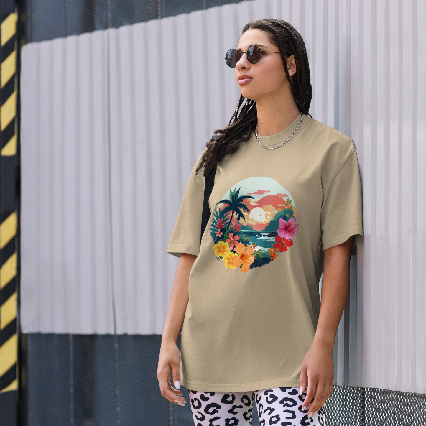 Fashion-forward woman in a Women's Tropical Sunset faded khaki Oversized Tee featuring a vibrant, artistic print of palm trees and flowers, paired with statement leopard print pants and chic sunglasses, encapsulating a fresh, summery aesthetic.