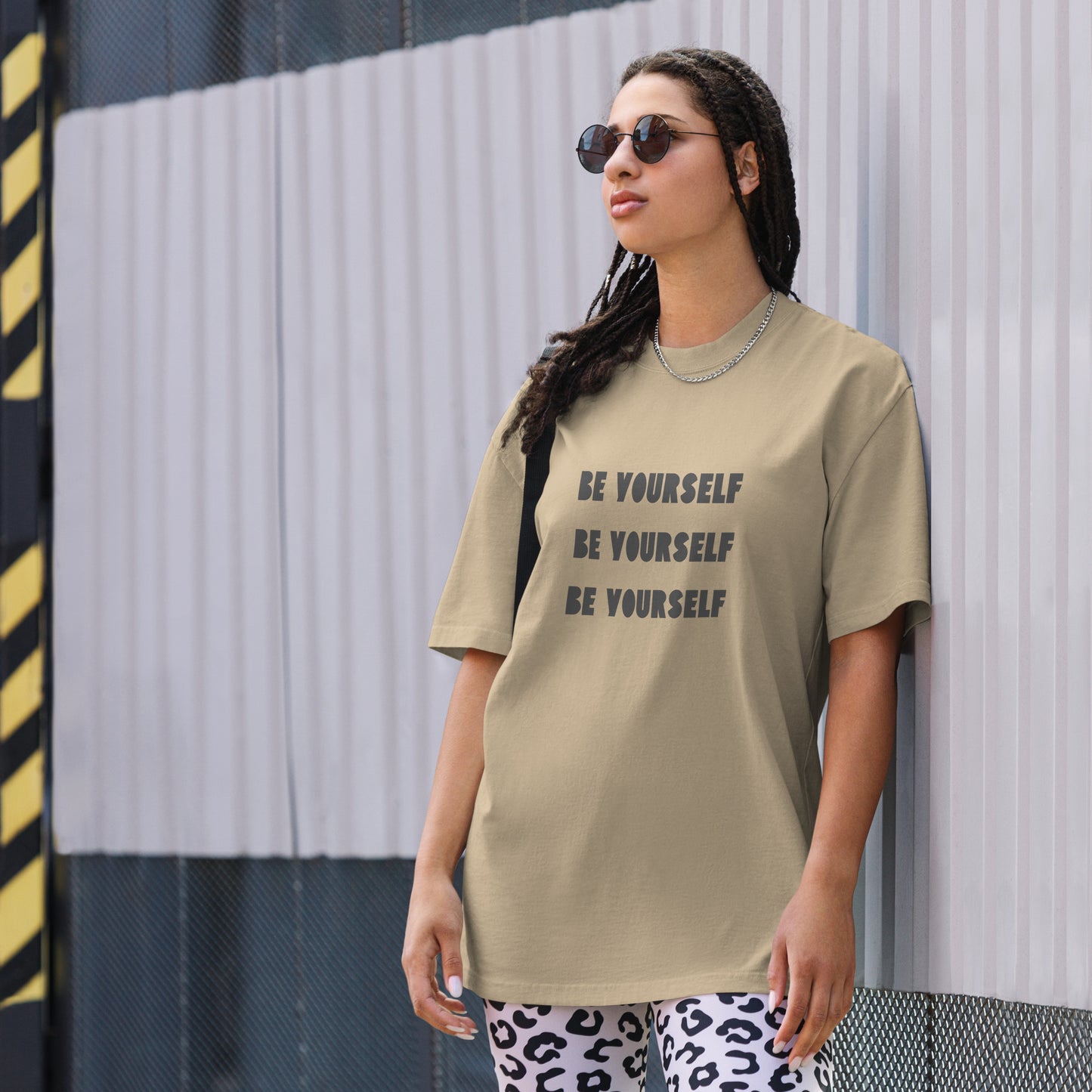 A self-assured woman wears the Women’s ‘Be Yourself’ faded khaki Oversized Tee leaning g against a wall wearing cheetah stretchy pants and circle sunglasses. 