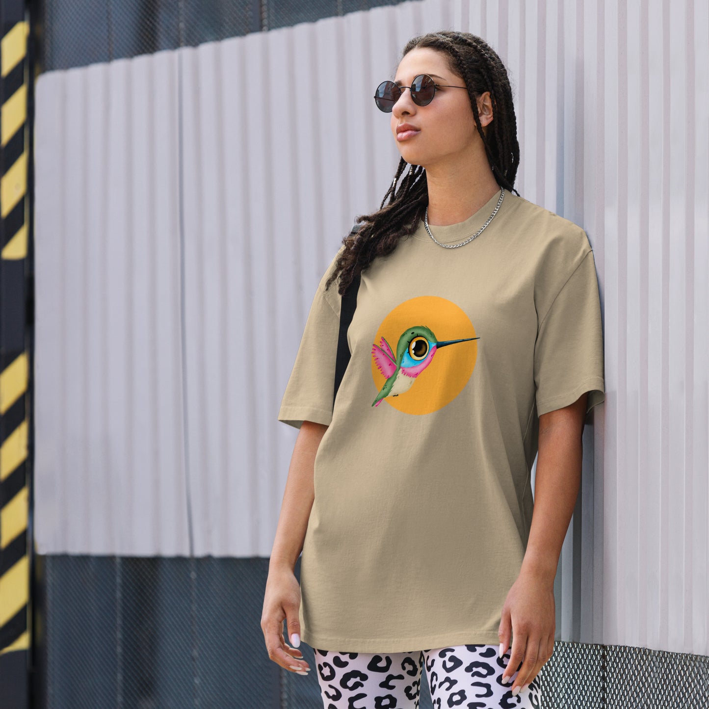 Woman models a faded khaki Oversized Tee with a colorful hummingbird design on a yellow circle background, embodying a lively spirit.