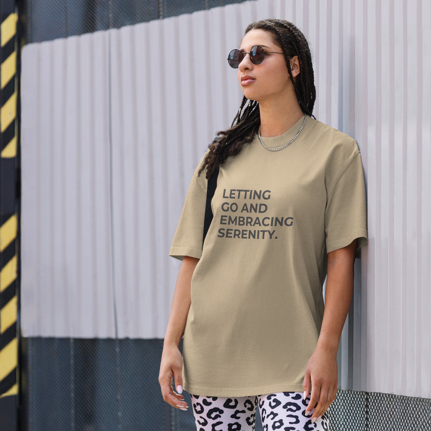 A woman wearing a faded khaki "Letting go and embracing serenity" t-shirt, styled with leopard print leggings and sunglasses, leaning against a corrugated metal wall with a confident expression. The casual yet trendy outfit showcases self-expression and boldness. Perfect for promoting empowering fashion statements.