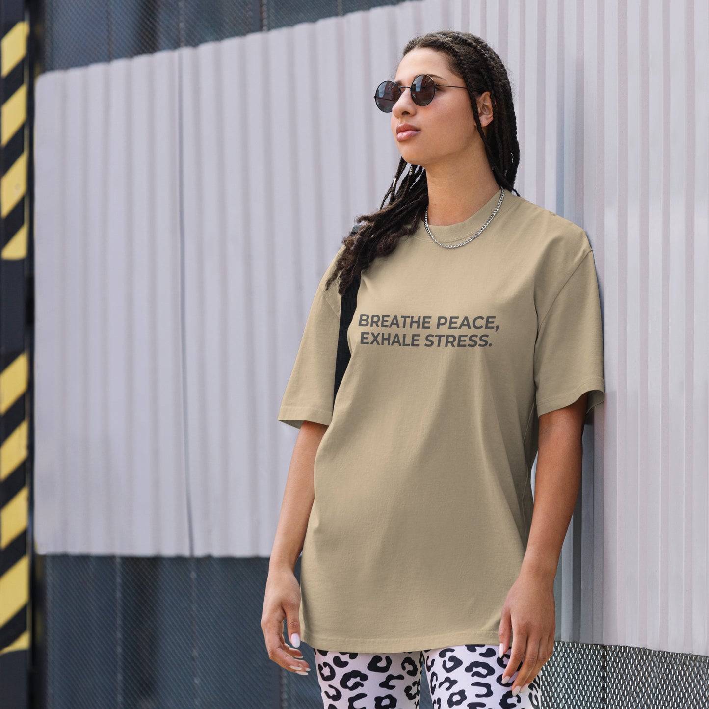 A woman wearing a faded khaki "Breath peace, exhale stress" t-shirt, styled with leopard print leggings and sunglasses, leaning against a corrugated metal wall with a confident expression. The casual yet trendy outfit showcases self-expression and boldness. Perfect for promoting empowering fashion statements.