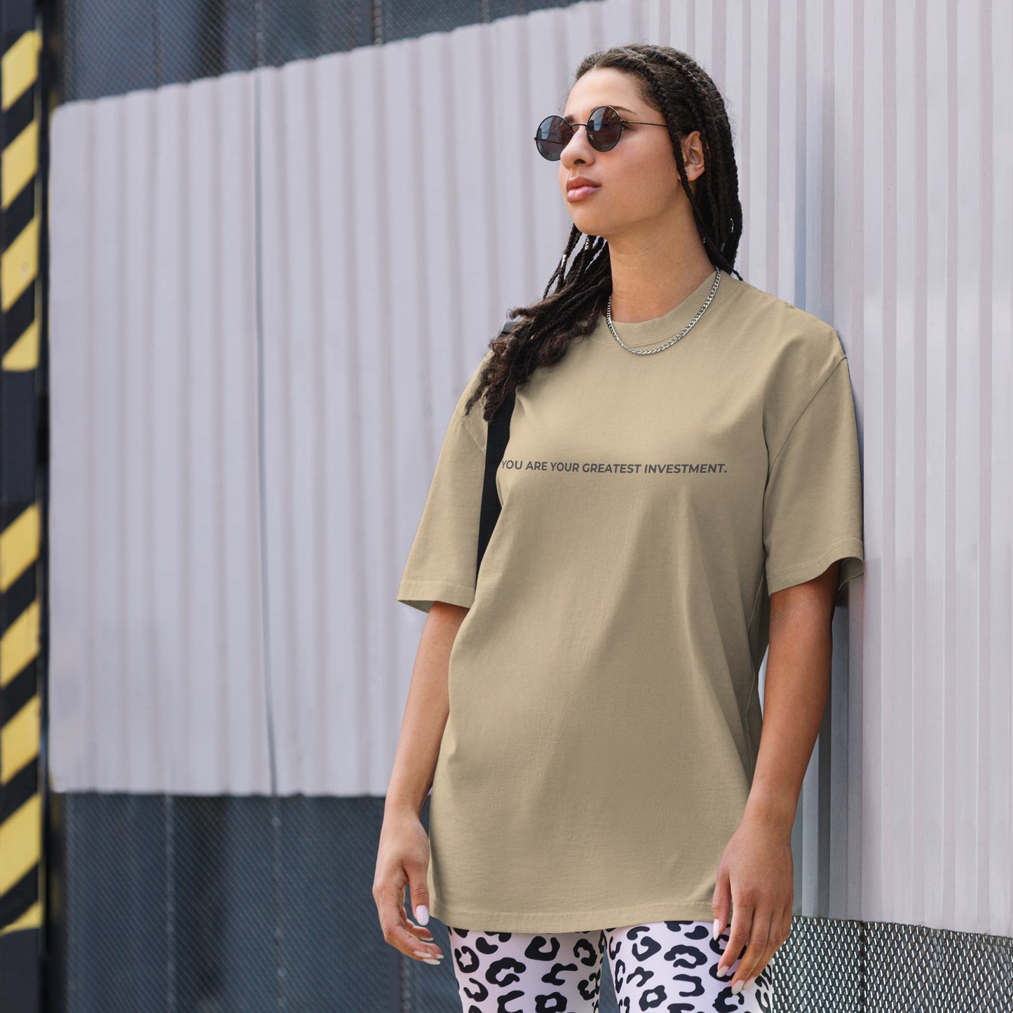 A woman wearing a faded khaki "You are your greatest investment" t-shirt, styled with leopard print leggings and sunglasses, leaning against a corrugated metal wall with a confident expression. The casual yet trendy outfit showcases self-expression and boldness. Perfect for promoting empowering fashion statements.