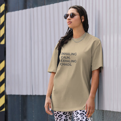 A woman wearing a faded khaki "Inhaling calm, exhaling chaos" t-shirt, styled with leopard print leggings and sunglasses, leaning against a corrugated metal wall with a confident expression. The casual yet trendy outfit showcases self-expression and boldness. Perfect for promoting empowering fashion statements.