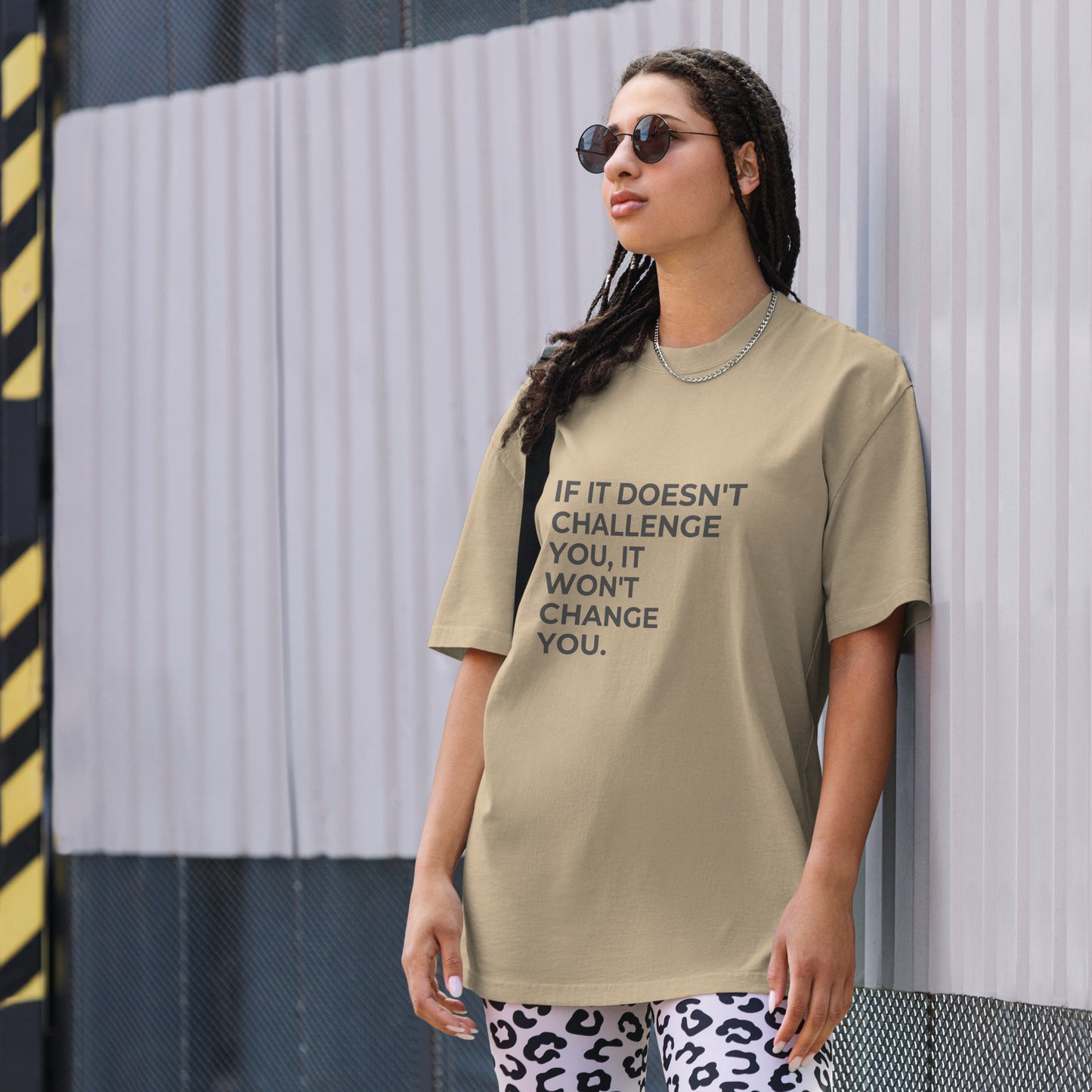 A woman wearing a faded khaki "If it doesn't challenge you, it won'f change you" t-shirt, styled with leopard print leggings and sunglasses, leaning against a corrugated metal wall with a confident expression. The casual yet trendy outfit showcases self-expression and boldness. Perfect for promoting empowering fashion statements.