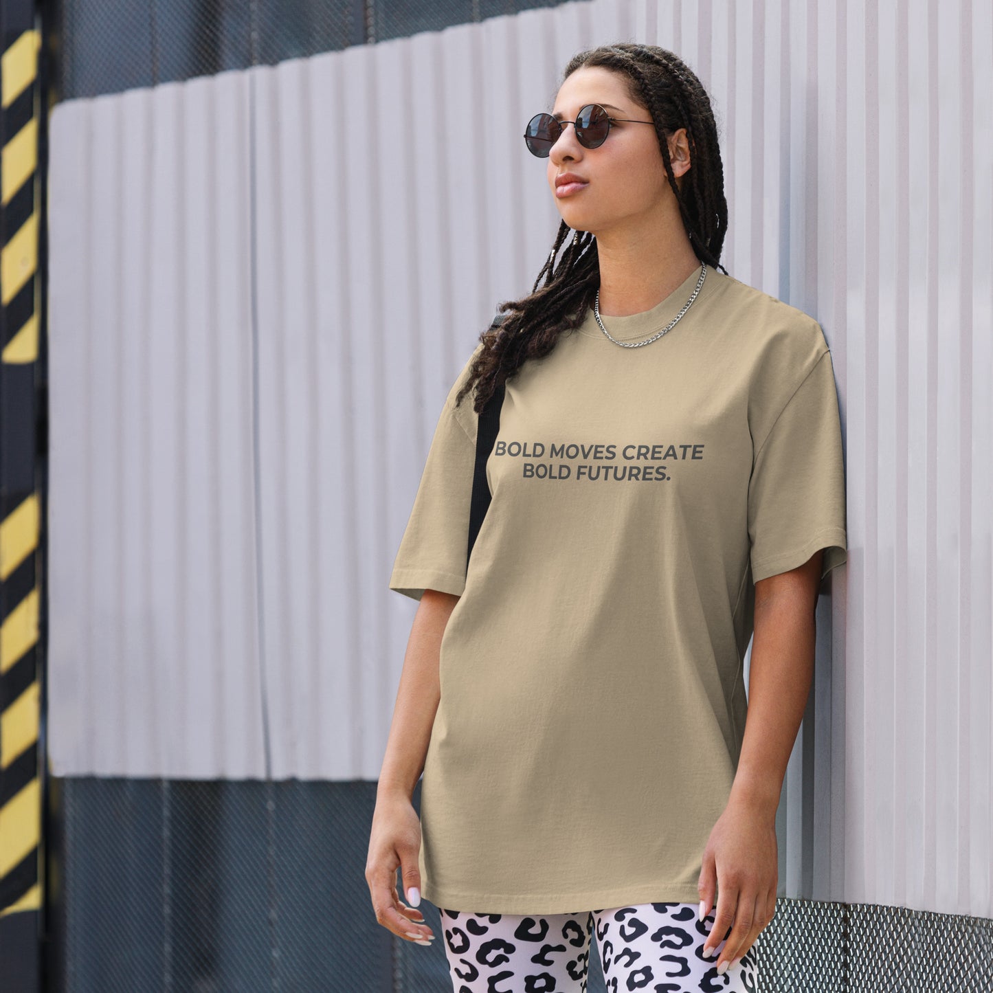 A woman wearing a faded khaki "Bold Moves Create Bold Futures" t-shirt, styled with leopard print leggings and sunglasses, leaning against a corrugated metal wall with a confident expression. The casual yet trendy outfit showcases self-expression and boldness. Perfect for promoting empowering fashion statements.