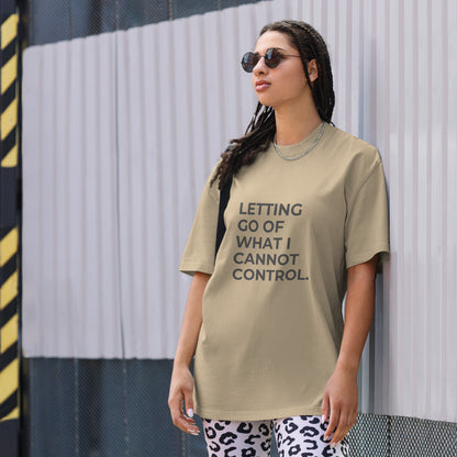 A woman wearing a faded kahki "Letting go of what I cannot control" t-shirt, styled with leopard print leggings and sunglasses, leaning against a corrugated metal wall with a confident expression. The casual yet trendy outfit showcases self-expression and boldness. Perfect for promoting empowering fashion statements.