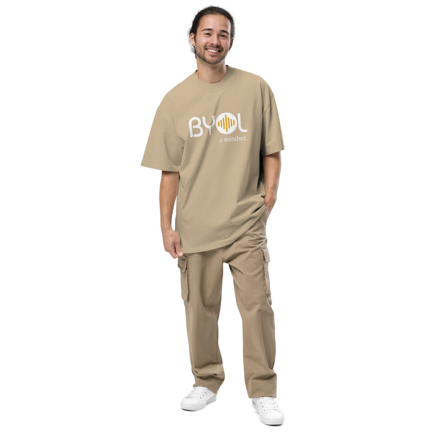 A young man wearing a khaki "BYOL: a mindset" T-shirt, smiling and standing with one hand in his pocket. The T-shirt features the "BYOL" logo in white and yellow on the front and is available in various colors (dark gray, beige, light blue) and sizes (S-3XL). The "be you out loud" logo is displayed at the top.