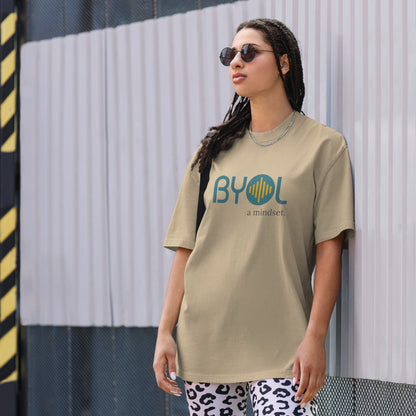 A young woman with long braids wearing sunglasses and a light faded khaki "BYOL: a mindset" T-shirt, standing and looking to the side. The T-shirt features the "BYOL" logo in teal and yellow on the front and is available in multiple colors (dark gray, beige, light blue, white, plus more) and sizes (S-3XL). The "be you out loud" logo is displayed at the top.