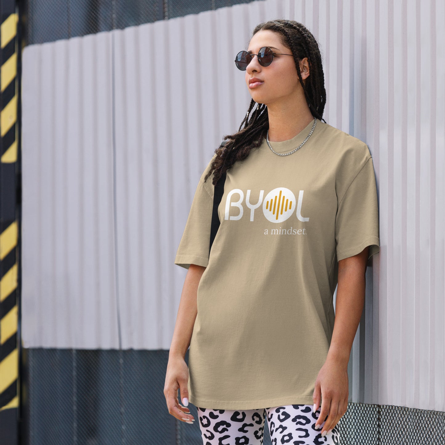 A young woman with long braids wearing sunglasses and a faded khaki "BYOL: a mindset" T-shirt, standing and looking to the side. The T-shirt features the "BYOL" logo in white and yellow on the front and is available in multiple colors (dark gray, beige, light blue, white, plus more) and sizes (S-3XL). The "be you out loud" logo is displayed at the top.