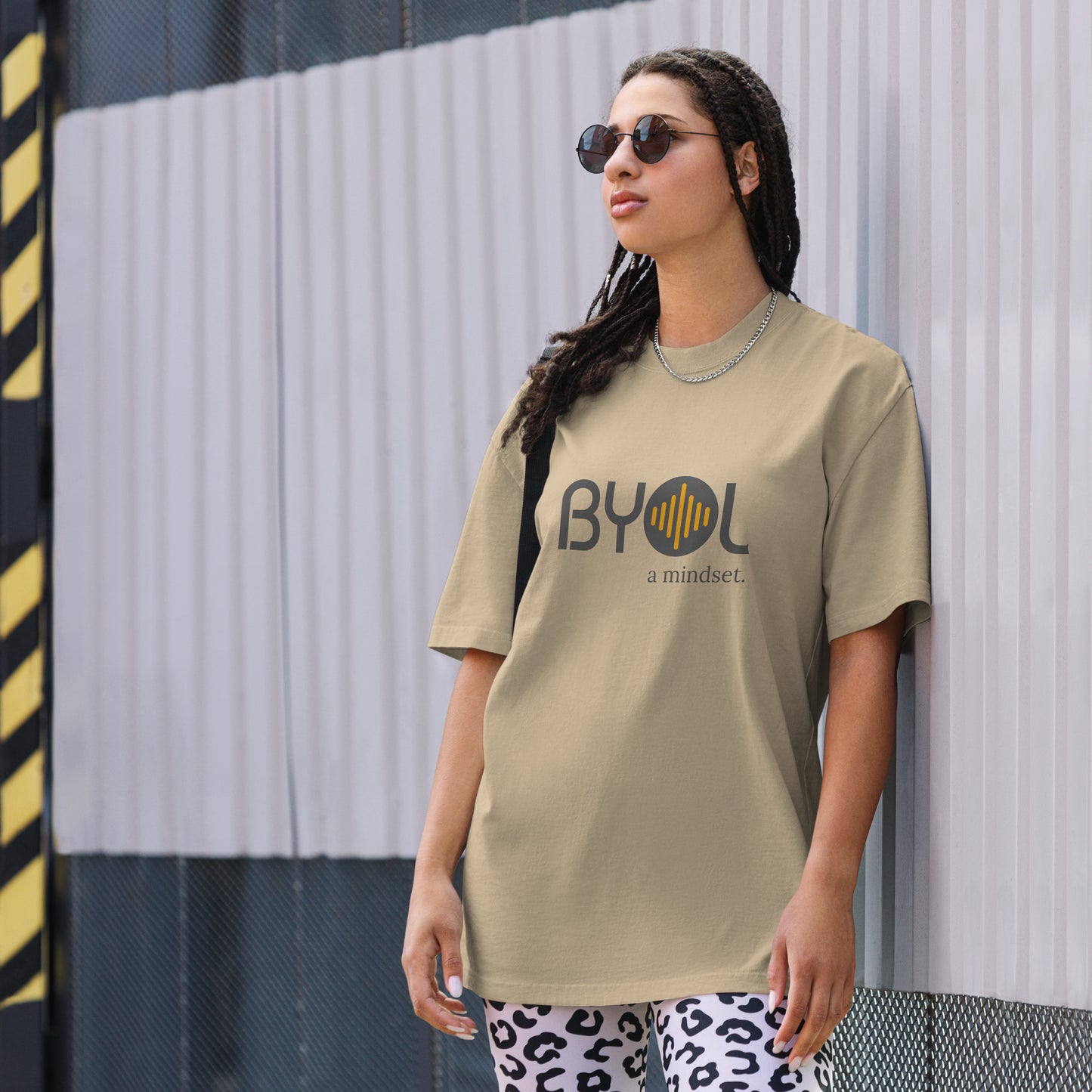 A young woman with long braids wearing sunglasses and a faded bone "BYOL: a mindset" T-shirt, standing and looking to the side. The T-shirt features the "BYOL" logo in dark gray and yellow on the front and is available in multiple colors (dark gray, beige, light blue, white, plus more) and sizes (S-3XL). The "be you out loud" logo is displayed at the top.