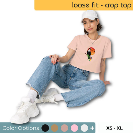 Relaxed woman in a pink crop top featuring a colorful toucan graphic, matched with loose denim jeans and casual white shoes showing the color and size options