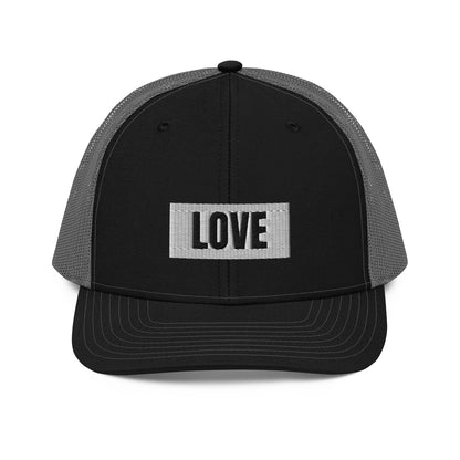 A black and grey snapback trucker cap with the word "LOVE" in white block letters inside a white rectangle, embroidered on the front panel. The hat features a curved brim, white mesh back panels for ventilation, and an adjustable snap closure for a comfortable, custom fit.