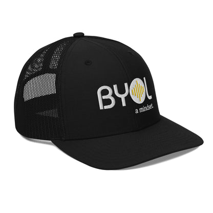 Black pre-curved contrast stitched trucker hat with "BYOL a mindset" embroidered on the front in white and yellow displayed against a clean background. Available in various colors: maroon, black, green, and gray.