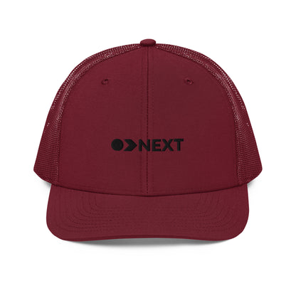 A maroon snapback trucker cap featuring a front panel with a black embroidered logo that reads "NEXT" with an eye-like symbol as the 'E.' The cap has a curved brim and breathable mesh panels on the sides and back, complete with a button top and adjustable strap for a versatile fit.