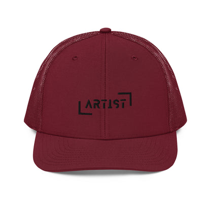 A maroon snapback trucker cap with the word "ARTIST" in bold black letters, framed by a black line square, embroidered on the front panel. It features a curved brim, button top, and black mesh back panels, offering a stylish look with a comfortable, breathable fit.