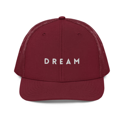 An maroon snapback trucker cap featuring the word "DREAM" in bold white letters embroidered across the front panel. This cap is designed with a curved visor, structured front crown, and breathable mesh panels on the sides and back, complete with an adjustable snap closure for a personalized fit.