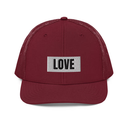 A maroon snapback trucker cap with the word "LOVE" in white block letters inside a white rectangle, embroidered on the front panel. The hat features a curved brim, white mesh back panels for ventilation, and an adjustable snap closure for a comfortable, custom fit.