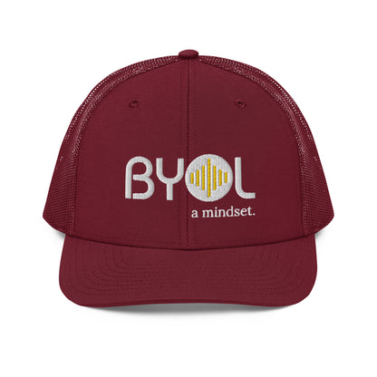 Maroon pre-curved contrast stitched trucker hat with "BYOL a mindset" embroidered on the front in white and yellow, displayed against a clean background. Available in various colors: maroon, black, green, and gray.