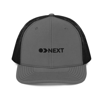 A grey and black snapback trucker cap featuring a front panel with a black embroidered logo that reads "NEXT" with an eye-like symbol as the 'E.' The cap has a curved brim and breathable mesh panels on the sides and back, complete with a button top and adjustable strap for a versatile fit.