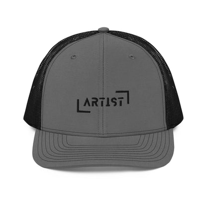 A charcoal gray snapback trucker cap with the word "ARTIST" in bold black letters, framed by a black line square, embroidered on the front panel. It features a curved brim, button top, and black mesh back panels, offering a stylish look with a comfortable, breathable fit.