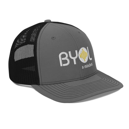 Grey and black pre-curved contrast stitched trucker hat with "BYOL a mindset" embroidered on the front in white and yellow displayed against a clean background. Available in various colors: maroon, black, green, and gray.