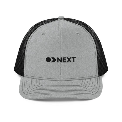 A grey and black snapback trucker cap featuring a front panel with a black embroidered logo that reads "NEXT" with an eye-like symbol as the 'E.' The cap has a curved brim and breathable mesh panels on the sides and back, complete with a button top and adjustable strap for a versatile fit.