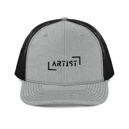 A grey and black snapback trucker cap with the word "ARTIST" in bold black letters, framed by a black line square, embroidered on the front panel. It features a curved brim, button top, and black mesh back panels, offering a stylish look with a comfortable, breathable fit.