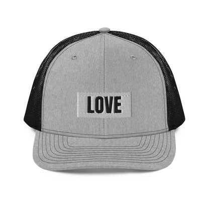 A grey and black snapback trucker cap with the word "LOVE" in white block letters inside a white rectangle, embroidered on the front panel. The hat features a curved brim, white mesh back panels for ventilation, and an adjustable snap closure for a comfortable, custom fit.