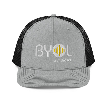 Grey and black pre-curved contrast stitched trucker hat with "BYOL a mindset" embroidered on the front in white and yellow, displayed against a clean background. Available in various colors: maroon, black, green, and gray.