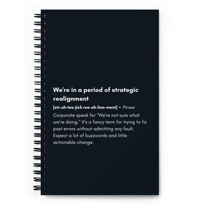 A spiral notebook with a black wire-o binding featuring a satirical definition of the corporate phrase "We're in a period of strategic realignment" printed in white text on a black background.