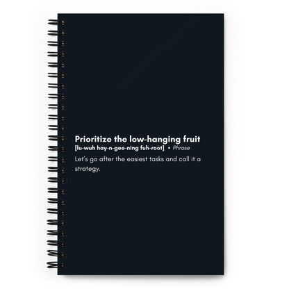 A black spiral-bound notebook with a satirical definition of the corporate phrase "Prioritize the low-hanging fruit" in white text on the cover, featuring a pronunciation guide and an explanation that humorously critiques the concept.