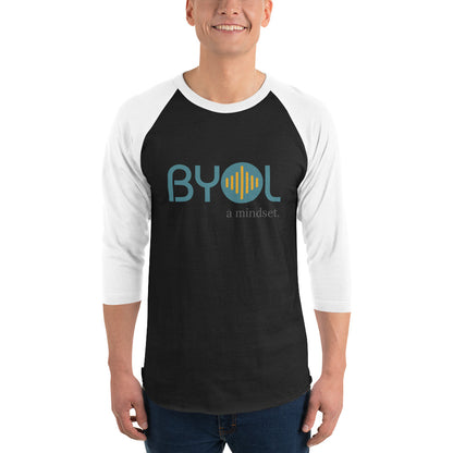 A young man wearing a black and white "BYOL: a mindset" raglan T-shirt with three-quarter sleeves, smiling and standing with his arms at his sides. The T-shirt features the "BYOL" logo in teal and yellow on the front and is available in multiple colors (red, black, white, green, plus more) and sizes (S-3XL). The "be you out loud" logo is displayed at the top.