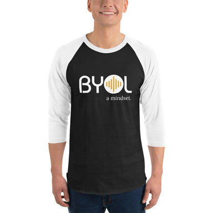 A young man wearing a black and white "BYOL: a mindset" raglan T-shirt with three-quarter sleeves, smiling and standing with his arms at his sides. The T-shirt features the "BYOL" logo in white and yellow on the front and is available in multiple colors (red, black, white, green, plus more) and sizes (S-3XL). The "be you out loud" logo is displayed at the top.