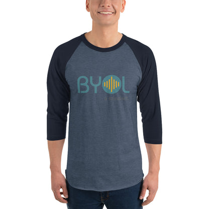 A young man wearing a heather blue and navy blue "BYOL: a mindset" raglan T-shirt with three-quarter sleeves, smiling and standing with his arms at his sides. The T-shirt features the "BYOL" logo in teal and yellow on the front and is available in multiple colors (red, black, white, green, plus more) and sizes (S-3XL). The "be you out loud" logo is displayed at the top.